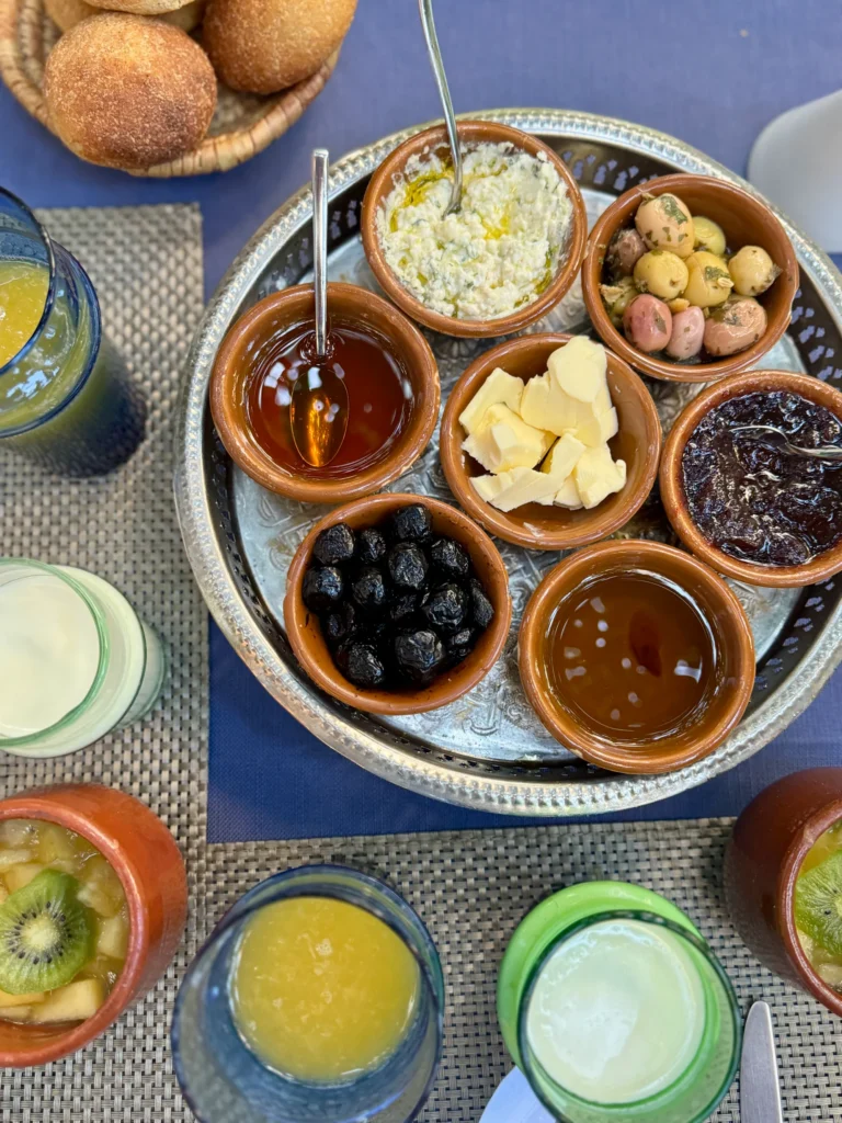 A tray with bowls of olives, butter, cheese, jam, honey, and olives, surrounded by drinks and bread on a blue tablecloth.