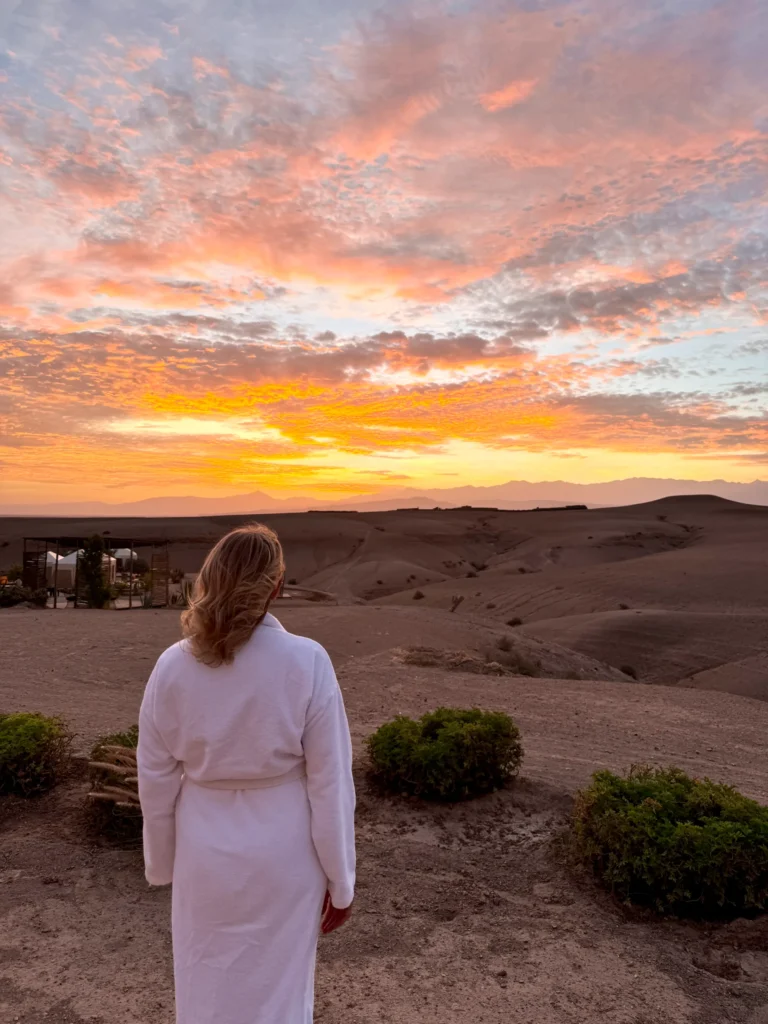 A person in a white robe stands on rocky terrain, facing a colorful sunset sky above a desert landscape with distant hills and small green bushes.