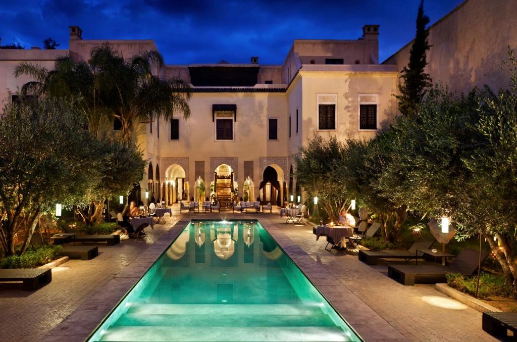 A rectangular illuminated pool is centered in a courtyard surrounded by trees, lounge chairs, and a building with arched doorways at night.