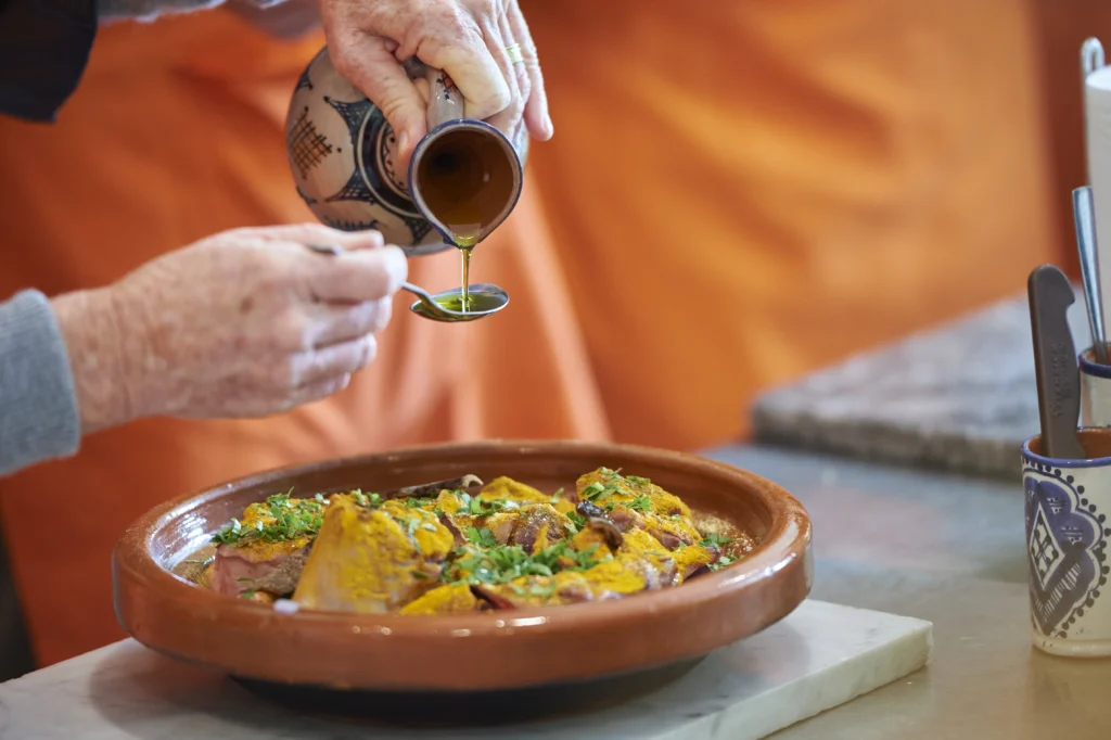A person pours liquid from a decorated jug onto a dish of seasoned meat, garnished with herbs, in a brown clay serving dish.