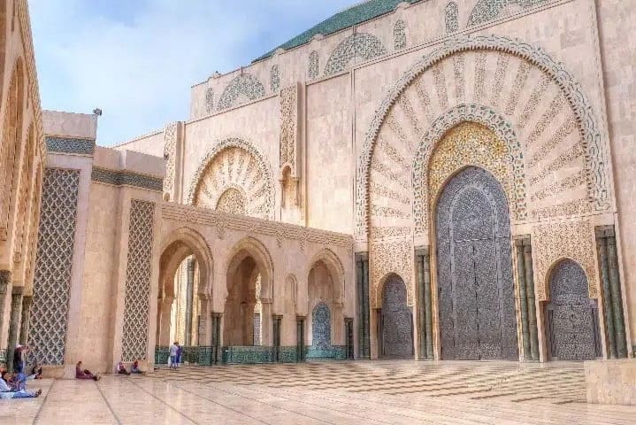 A large mosque courtyard with ornate archways, intricate tilework, and a grand decorated entrance door. People are sitting along the edges.