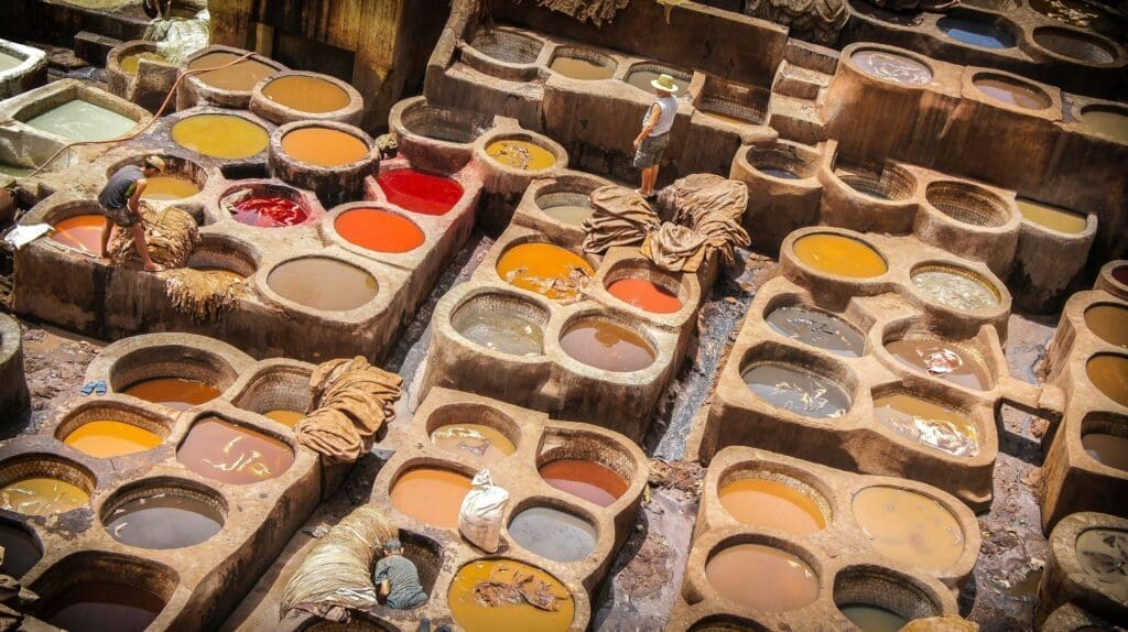 Workers stand among large, colorful vats filled with dyes and hides at a traditional outdoor tannery. The vats are arranged in an irregular pattern.