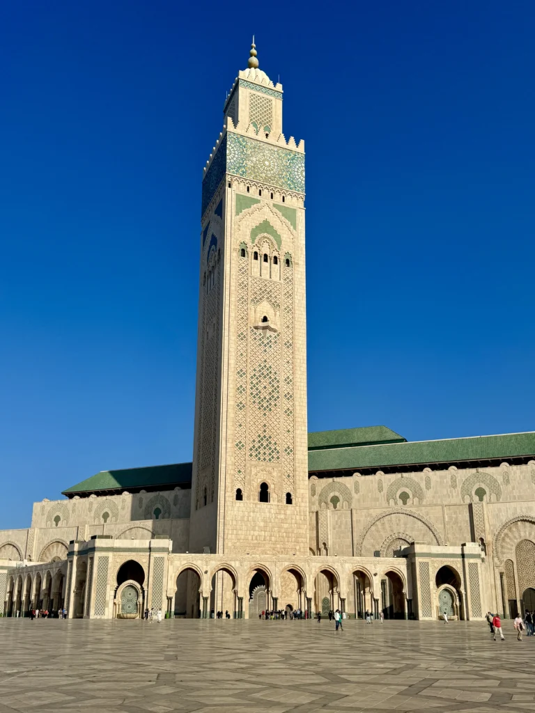The Hassan II Mosque in Casablanca, Morocco, with its tall minaret and detailed exterior, stands under a clear blue sky with people visible in the courtyard.
