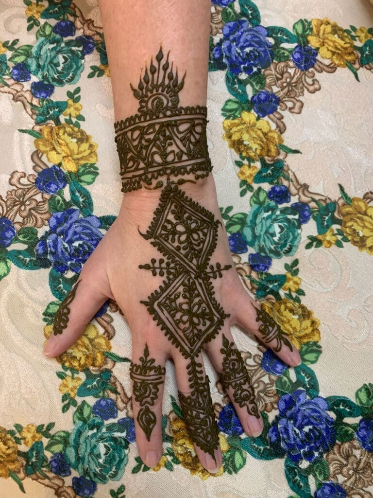 A hand with intricate henna designs covering the back and fingers, displayed on a floral-patterned fabric.