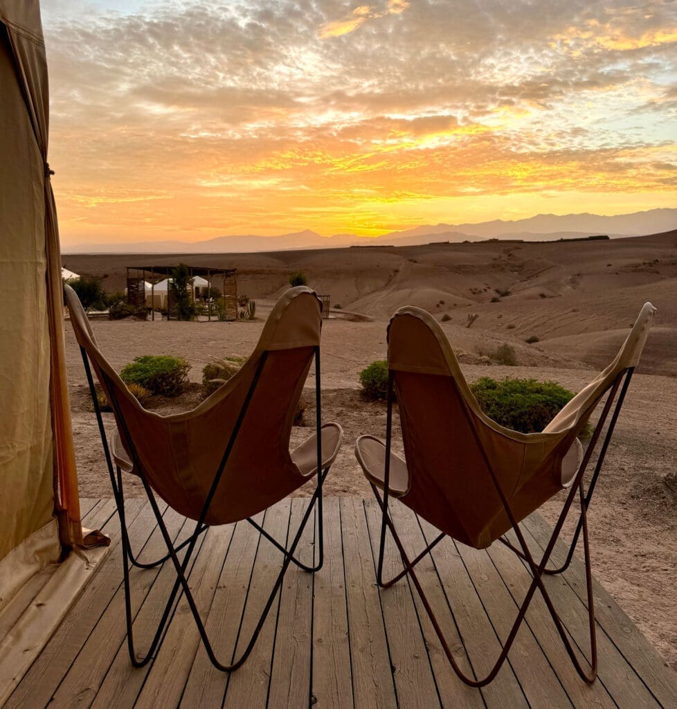 Two empty canvas chairs on a wooden deck overlook a desert landscape at sunset, with scattered clouds and distant mountains.