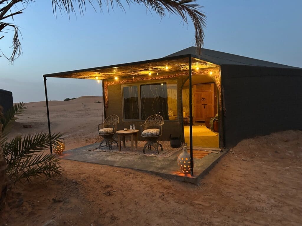 A desert tent with a lit patio, two wicker chairs, a table with water bottles, decorative lanterns, and sand dunes in the background at dusk.