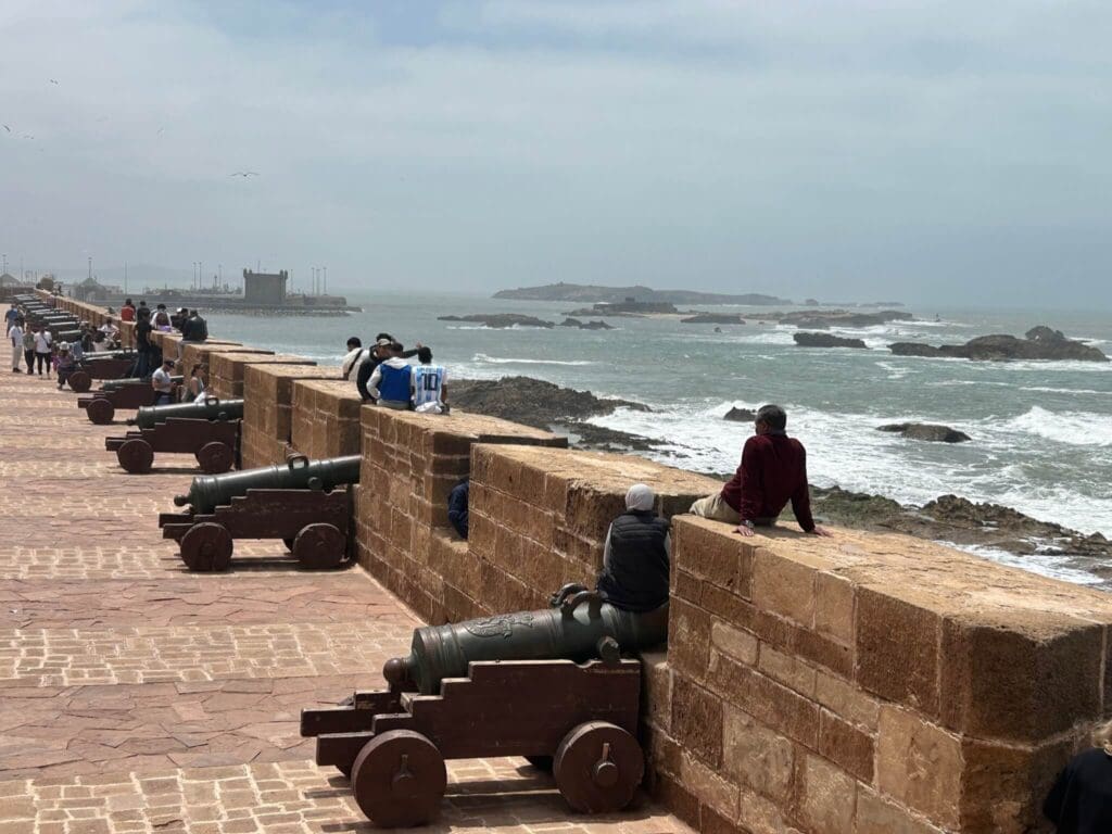 People sit and walk along a stone coastal fortification lined with old cannons, overlooking a rocky shoreline and the ocean under a cloudy sky—a perfect scene for exploring Morocco travel trade or forming B2B partnerships in an inspiring setting.