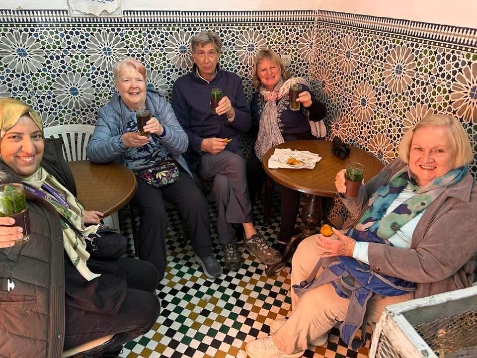 Five people sit around two small tables with mosaic tile walls and floors, holding green drinks and smiling at the camera in a cozy, brightly patterned room—perfect for inspiring B2B travel partnerships with Morocco Travel Organizer.