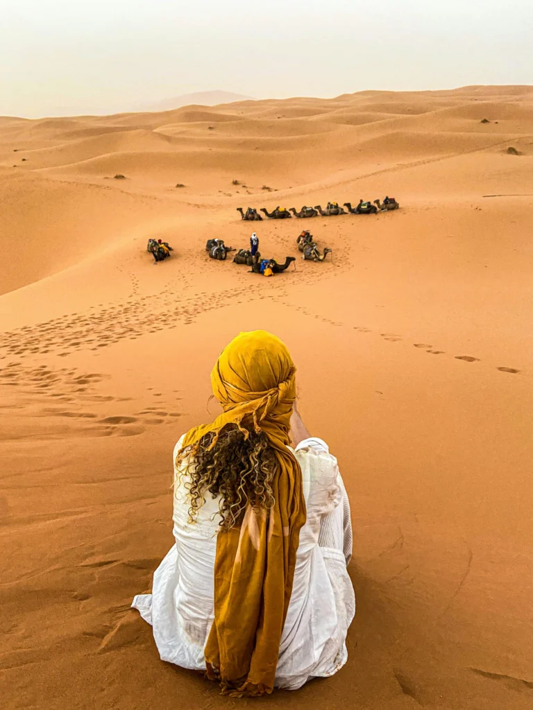 A woman sitting in the desert looking at camels.