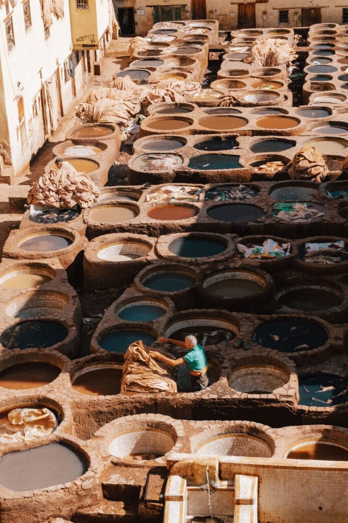 A worker stands among large stone vats filled with various colored liquids at a traditional tannery, surrounded by buildings in sunlight.