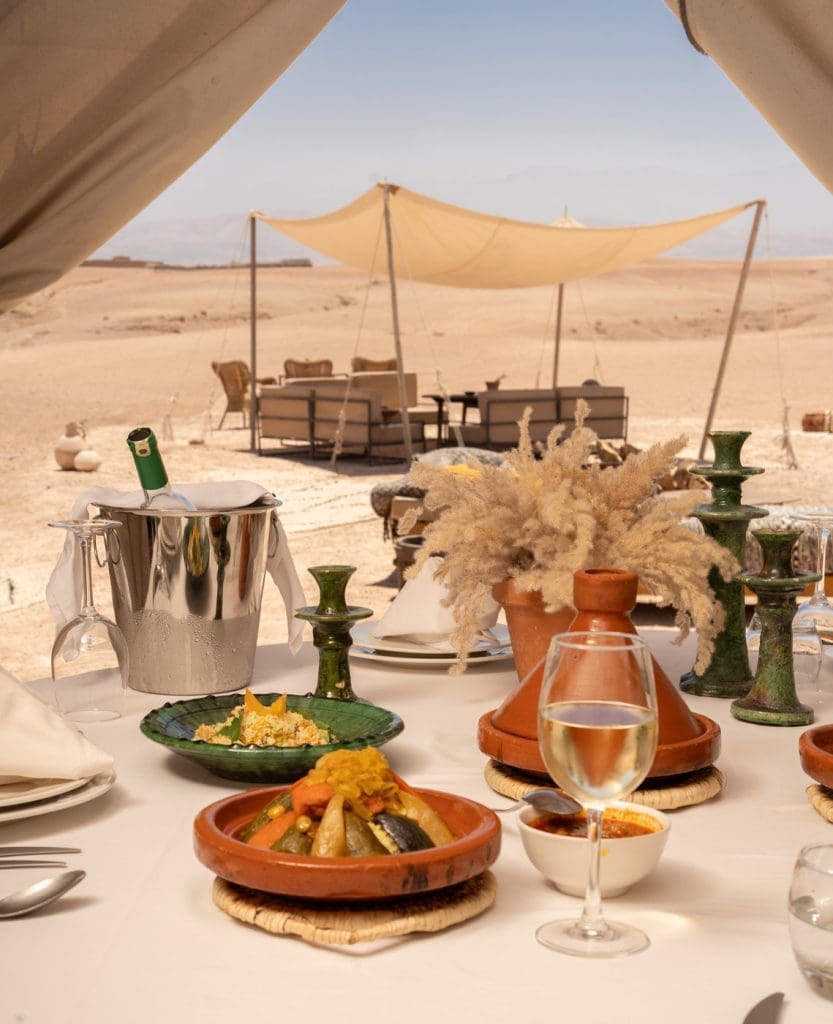 A table set with food, wine, and green candles under a tent overlooks a lounge area and desert landscape with sandy terrain and a canopy in the background.