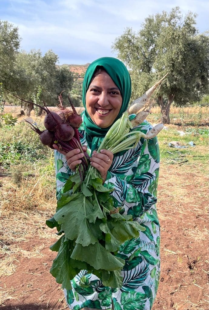 A woman in a green patterned dress and headscarf smiles while holding fresh beets and white radishes in a rural garden setting with trees in the background.