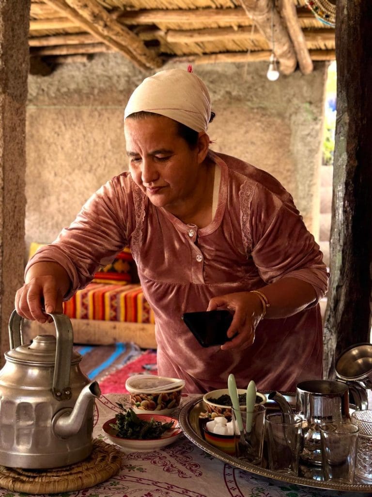 A woman in a pink dress and headscarf pours tea from a metal teapot at a table set with dishes, glasses, and snacks in a rustic, indoor setting.